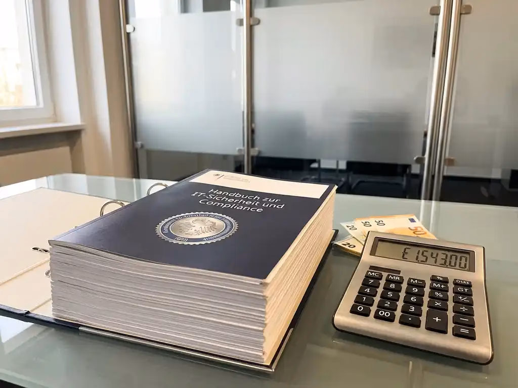 Deutsches BSI-Compliance-Handbuch aufgeschlagen auf Bürotisch neben Taschenrechner und Euro-Banknoten im modernen Büro