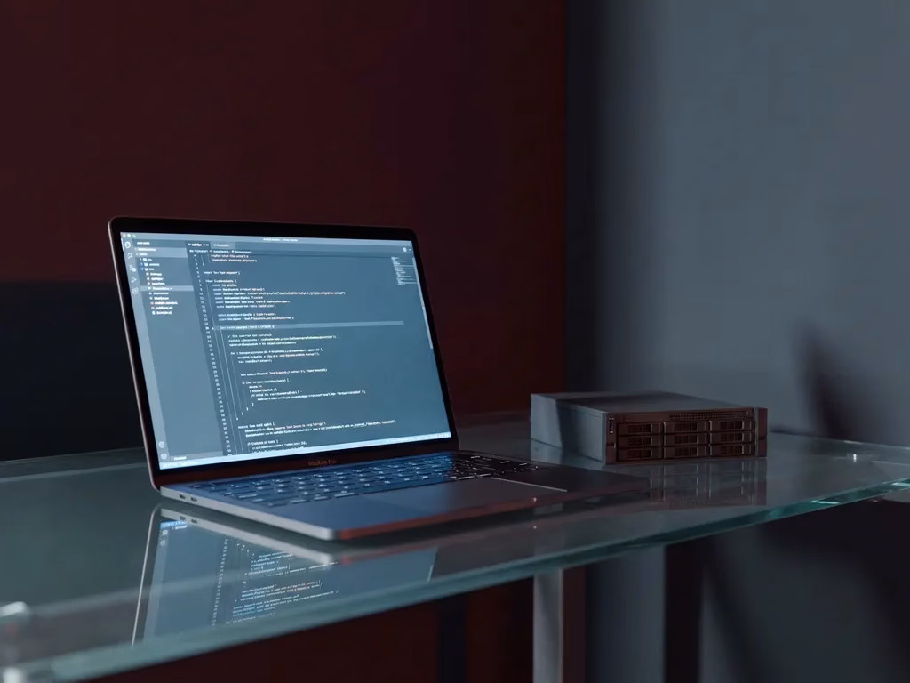 Laptop eines Red-Team-Analysten auf Glasschreibtisch, daneben ein kleines Servermodell, stahlblauem Umgebungslicht und dunklem Hintergrund.