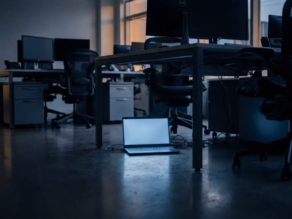 Nicht autorisiertes Laptop leuchtet schwach unter einem Schreibtisch in einem dunklen Büro mit blauem Lichtschein auf Betonboden.